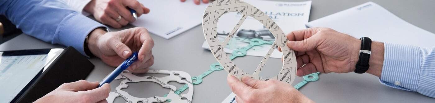 3 men at a desk. The first man on the left is sat infront of an ipad and he is holding a KLINGER pen. The man next to him is taking notes. The man on the right is holding gasket sheeting material, and on the table there are more gasket sheeting materials with an installation guide