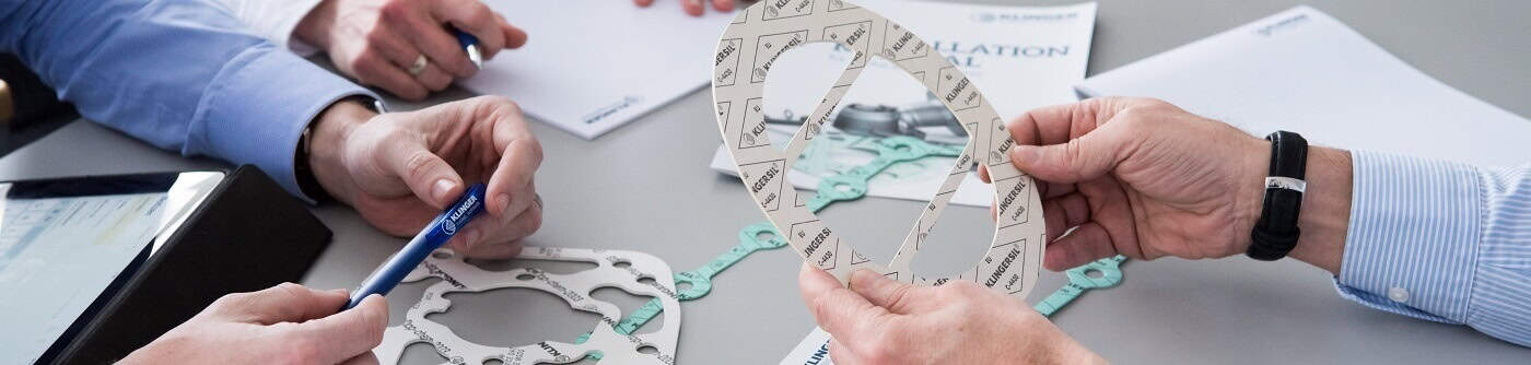 3 men at a desk. The first man on the left is sat infront of an ipad and he is holding a KLINGER pen. The man next to him is taking notes. The man on the right is holding gasket sheeting material, and on the table there are more gasket sheeting materials with an installation guide