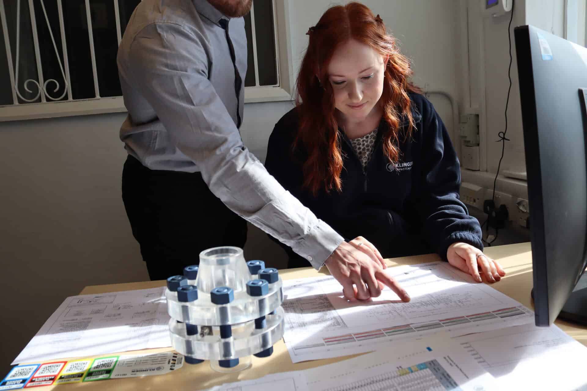 female KLINGER employee being instructed by a fellow employee of flange management map, on a desk with flange displayed beside images, computer on far right and flange tags on the far left.