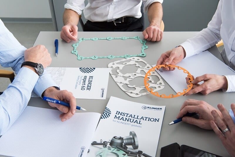 Point of View style image of KLINGER employees in a meeting at a table presented with KLINGER EXPERT documentation and manuals along with cut sheeting materials